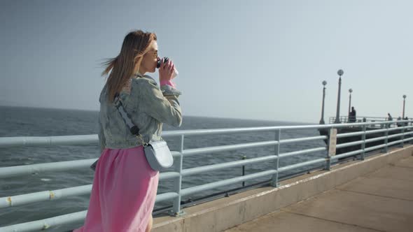 Stylish Happy Woman in Flowing Pink Dress Walking By Ocean Pier Drinking Coffee alt