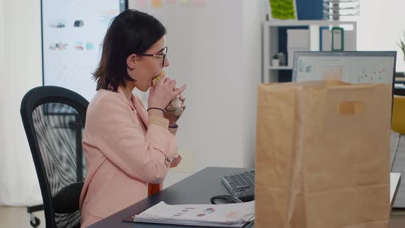 Businesswoman Eating Bite of Tasty Sandwich Drinking Coffee in Front of Monitor alt