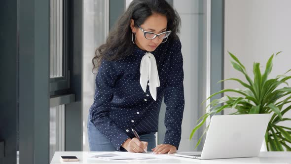 Young mixed race woman entrepreneur using laptop writing notes stand at office desk