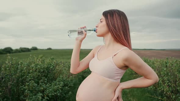 Pregnant young woman with dark hair in a pink top and light pants drinks water outdoors.