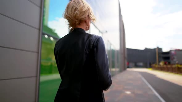 the Blonde Goes Along the Wall with the Big Windows, Turns Around and Takes Off Her Black Jacket alt