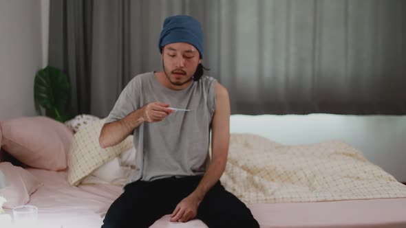 Asian man with cold checking temperature with a thermometer while in the bedroom at home.