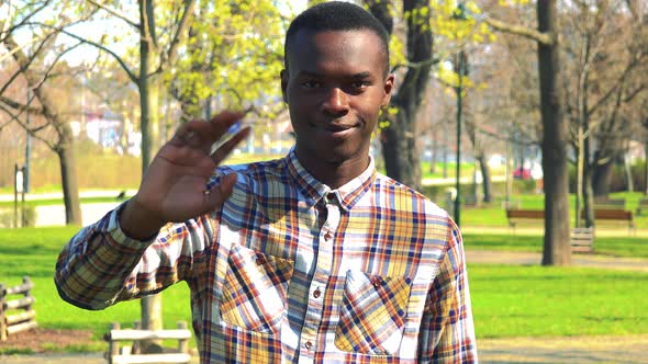A Young Black Man Smiles and Waves at the Camera in a Park on a Sunny Day alt