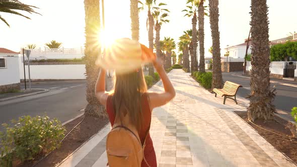 Happy Woman in a Big Yellow Hat Walking Along a Palm Alley at Sunset alt