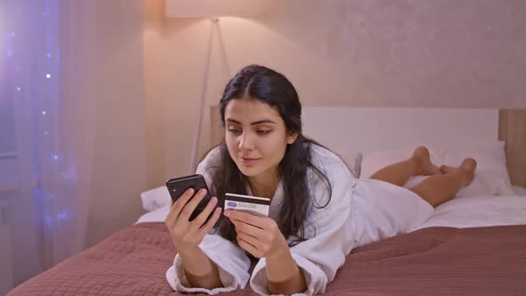 Smiling Young Woman Customer Holding Credit Card and Smartphone Sitting on Couch at Home alt
