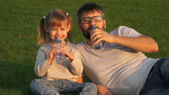 Father with Little Child Smile Laugh and Drink Water From Bottles Sitting on Lawn in Park