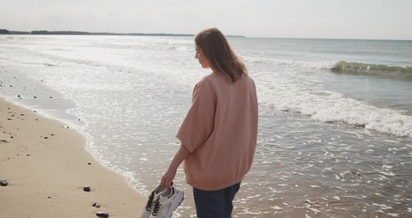 Young Attractive Woman Walking Along Beach Holding Sneakers in the Hand