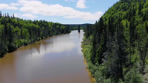A drone flies above the water of a river with trees and mountains on both sides and an old railroad alt