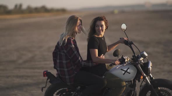 Attractive Young Woman Motorcyclist with His Girlfriend Riding a Motorcycle in a Desert on Sunset or alt