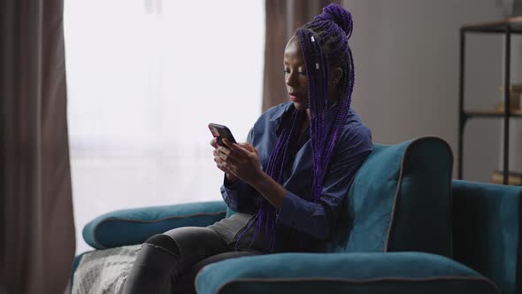 Afroamerican Lady with Modern Smartphone is Resting at Home Sitting Alone on Couch and Reading News alt