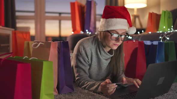 Woman with Glasses Wearing a Santa Claus Hat is Lying on the Carpet and Makes an Online Purchase alt
