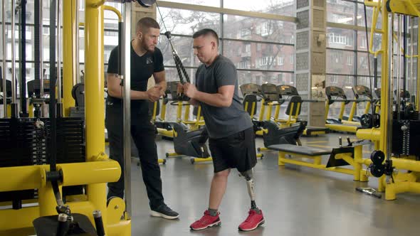 Disabled Young Man Working Out with the Help of Instructor in Gym