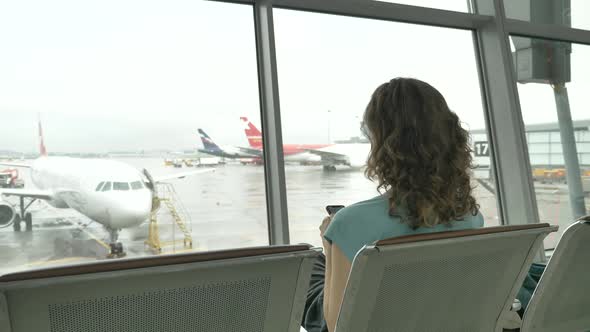 A Young Woman is Sitting with a Phone at the Airport in the Waiting Room Rear View alt
