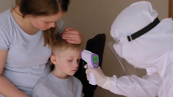 A Doctor in a Protective Suit Measures the Temperature to a Small Boy at Home alt