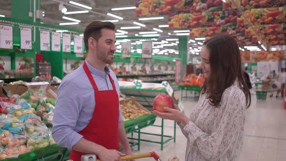 Friendly handsome male stock clerk in red apron, helping young attractive woman to choose fruits 