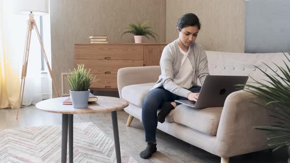 Pretty young lady sitting on couch at home, using laptop, watching videos online, chatting