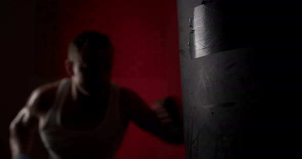 A man in a semi-dark hall, wearing hand wraps, hits a punching bag with quick and accurate punches. alt