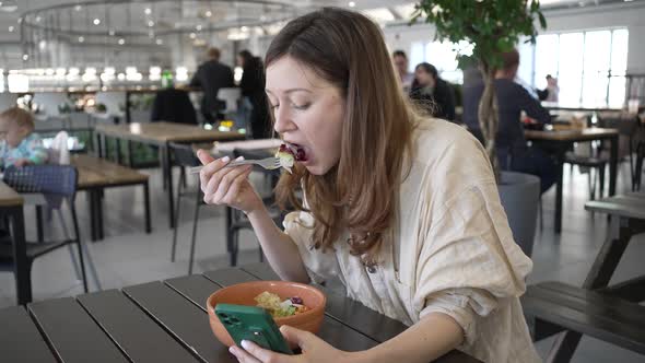 Woman in Cafe Eats Salad Scrolling Social Media Pages alt