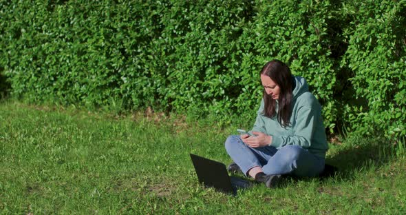 Woman Is Working Using Smartphone And Laptop Sitting On Grass In Park