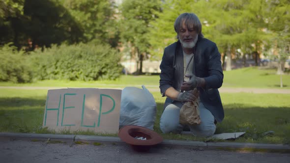 Homeless Old Beggar Asking for Money Sitting on Ground Outdoors and Drinking Alcohol alt