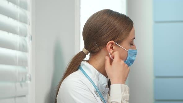 Exhausted young woman in white coat with stethoscope on neck takes off blue disposable face mask alt