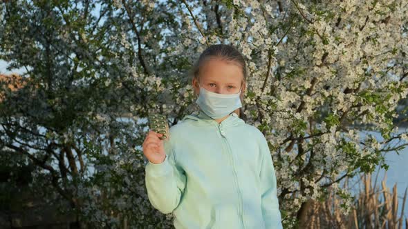 Allergy pills. A Little girl by a flowering tree in a medical mask.