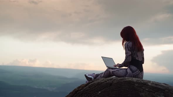 Girl Blogger Traveler is Typing Text on Laptop While Sitting on Top Mountain alt