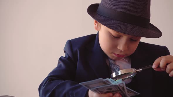 Child Boy Looks Like a Businessman in Hat and Suit is Looking at Dollars with Magnifier alt
