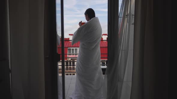 Woman in a White Blanket Stands in the Doorway with a Cup of Tea alt
