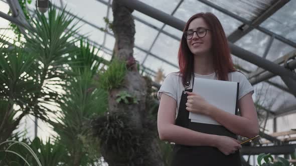 Happy Young Woman Scientist or Botanist Stands in a Greenhouse