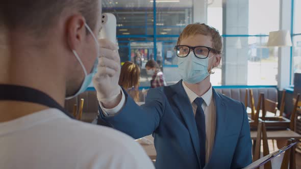 Young Manager in Protective Mask and Gloves Check Temperature on Waiter Forehead. alt