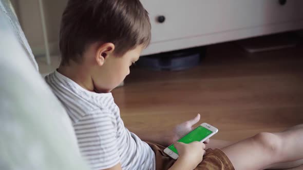 small boy sitting on floor and watching content on phone. Green screen mock-up.