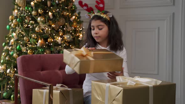 Girl is Sitting on a Chair Next to a Christmas Tree in a Christmas Hat with Reindeer Ears and alt