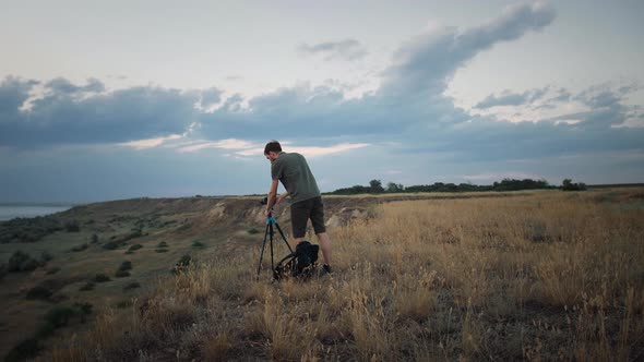 Guy Photographer Placing Camera Fixed on Tripod on Ground of High Hill and Adjusting It alt