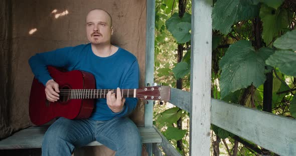 Man with an Acoustic Guitar is Sitting on Porch of House Playing an Instrument alt