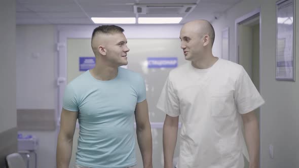 Two Positive Caucasian Men Walking Along Hospital Corridor and Talking. Portrait of Happy Patient alt