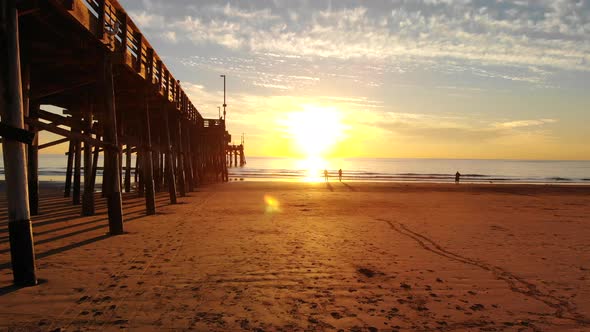 Drone flying under Newport Beach pier at sunset as sunlight beams and people walk the California coa alt