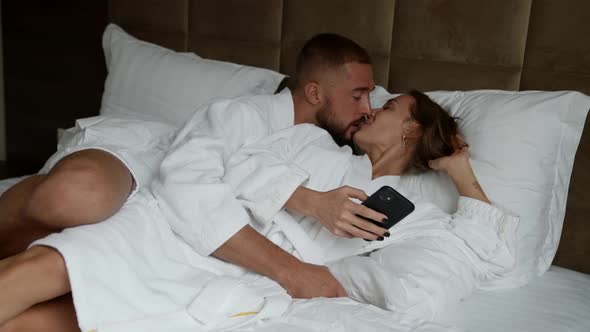Morning in Hotel Room Man and Woman are Embracing in Bed Lady is Using Smartphone alt