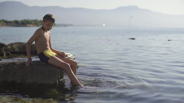 A Teenager Is Sitting on a Large Rock and Soaking His Feet in Water. The Boy Holds a Diving Mask in