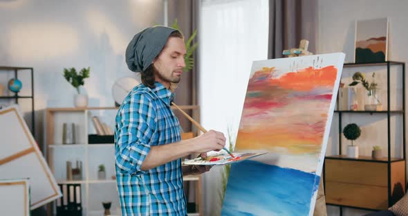 Male Artist Standing Near Easel with Big Canvas and Drawing Picture in Home Studio alt