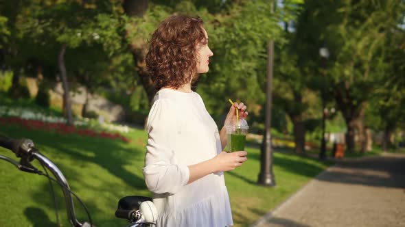 Attractive Young Woman Standing in the City Park Near Her City Bicycle with Flowers in Its Basket alt