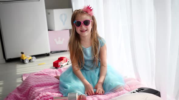 Little girl in a beautiful blue dress, a crown and sunglasses laughs looking at the camera.