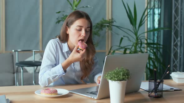 Young Businesswoman Eating a Glazed Donut While Working on a Laptop at the Office Table alt