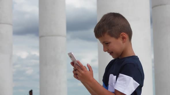 Tanned Boy is Watching at Phone in His Hands on the Background of White Columns alt