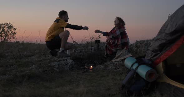 Two Young Travelers Man and Woman Sitting Near the Campfire and Tent Outdoors During Sunset and