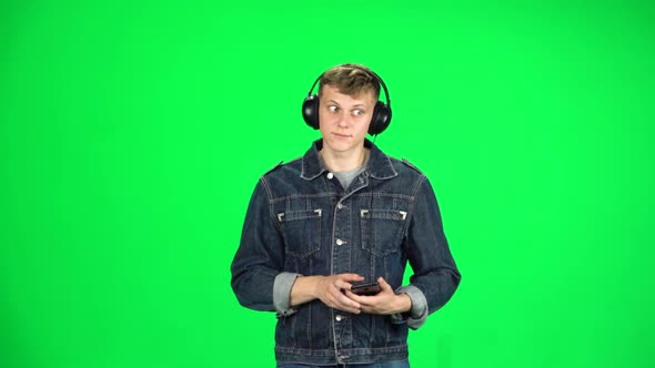 Guy in Big Headphones Goes, Listening Music and Dancing with Smartphone on Green Screen at Studio alt