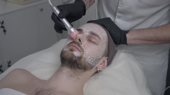 Face of Caucasian Man with Closed Eyes with Beautician Cleansing Nose Skin with Red LED Light Device alt