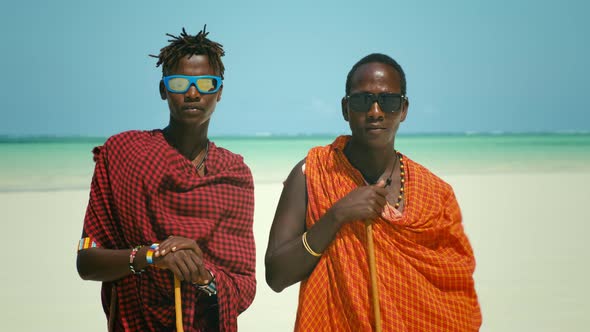 Young Masai Tribe Members in Red Dresses and Sunglasses Standing on Sand Beach of the Indian Ocean