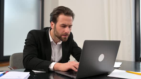 Puzzled Young Businessman Sitting at Desk in Office and Watching at Laptop Screen