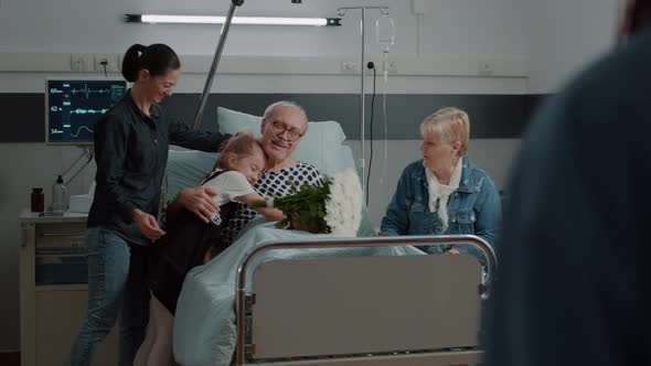Family Visiting Old Patient and Bringing Flowers in Hospital Ward Bed alt